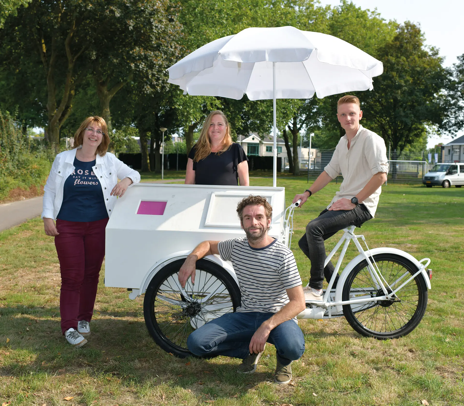 Vier mensen bij een witte bakfiets met parasol in een groene woonomgeving.